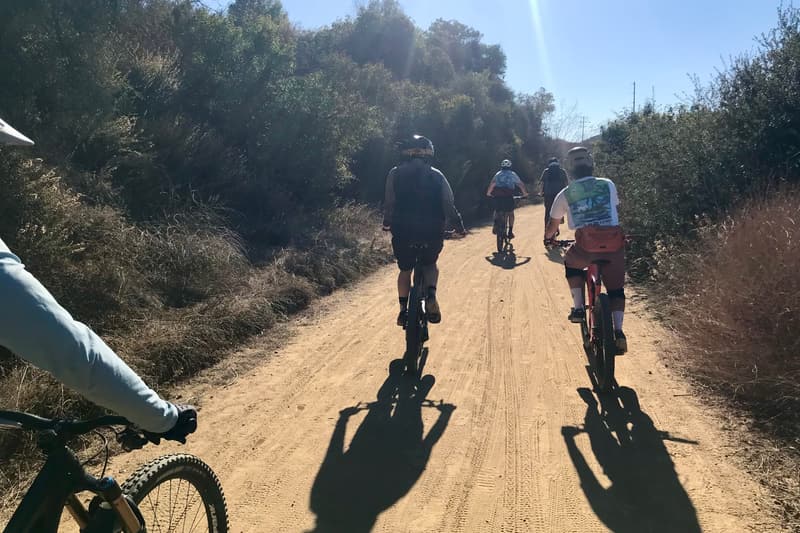 A crew of five riding single file up a dusty fire road at midday, photographed from the back of a sixth bike with its handlebars in frame.